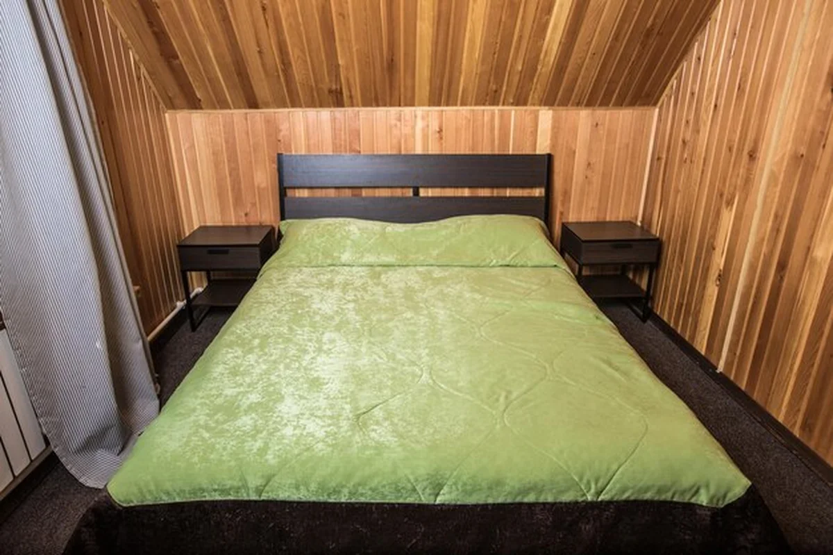 Podwójne łóżko z zielonym nakryciem w hotelowej sypialni z drewnianymi ścianami, oknem i zasłoną